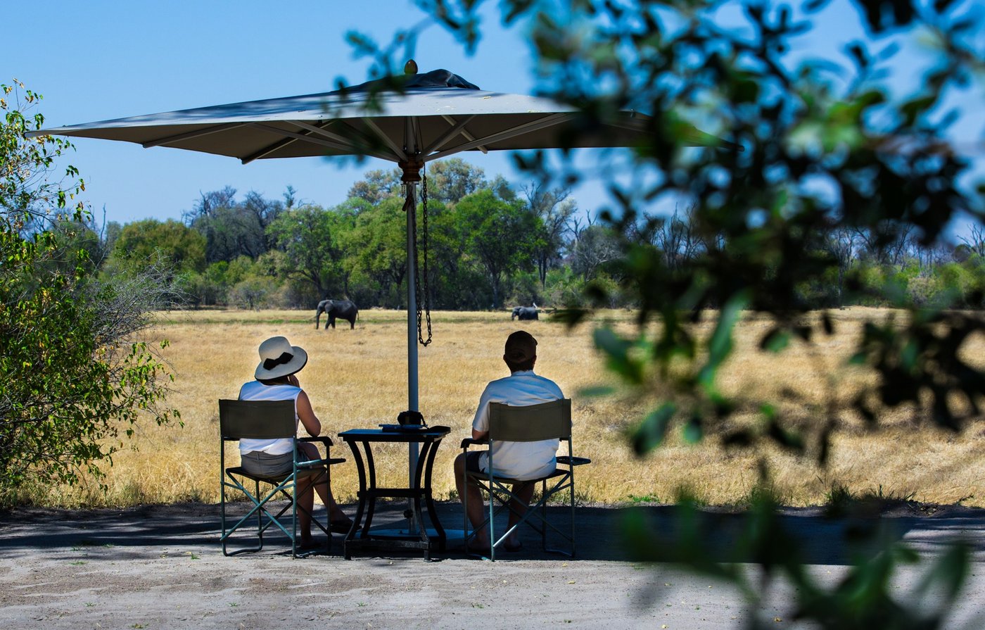 Dining deck under an umbrella with an elephant grazing beyond at Mana River Camp.